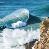 A large ocean wave crashes near a rocky cliff with a lighthouse, surrounded by onlookers and surfers in the water.