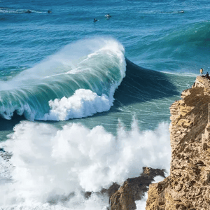 A large ocean wave crashes near a rocky cliff with a lighthouse, surrounded by onlookers and surfers in the water.