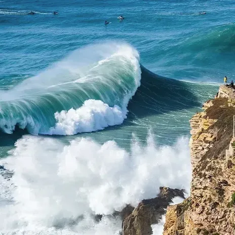 A large ocean wave crashes near a rocky cliff with a lighthouse, surrounded by onlookers and surfers in the water.