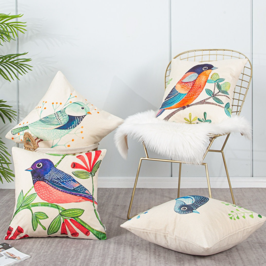 Decorative pillows with colorful bird designs are arranged on a metal chair with a faux fur rug. A plant and magazines are nearby, creating a cozy setting.