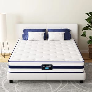 A white, tufted mattress labeled \“NISIEN\“ with blue accents sits on a white bed frame. It is flanked by two blue and white pillows, a plant, and a floor lamp, on a patterned gray rug.