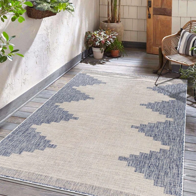 A 4'3\“ x 5'11\“ Southwestern outdoor rug with a geometric pattern in neutral tones, set on a wooden deck with surrounding plants and a chair with a striped cushion.