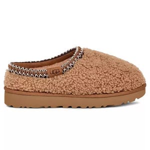 A brown, cozy UGG slipper with a textured, fluffy exterior and a cushioned sole, featuring embroidered trim around the opening.