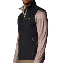 A man models a Columbia Sportswear zip-up black vest over a long-sleeve beige shirt, featuring a camouflage pattern on the inside collar.
