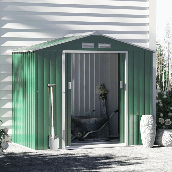 Green metal garden shed with white doors, a shovel, wheelbarrow, and decorated with two white ornamental vases.