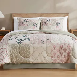 A quilted patchwork bedding set with floral and geometric patterns in shades of pink, beige, and green is displayed on a neatly made bed, accompanied by two matching pillowcases.