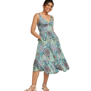 A woman is wearing a sleeveless, knee-length, floral-patterned dress with a sweetheart neckline and a ruffled hem. She has on white strappy sandals and is smiling with her hands in her pockets.