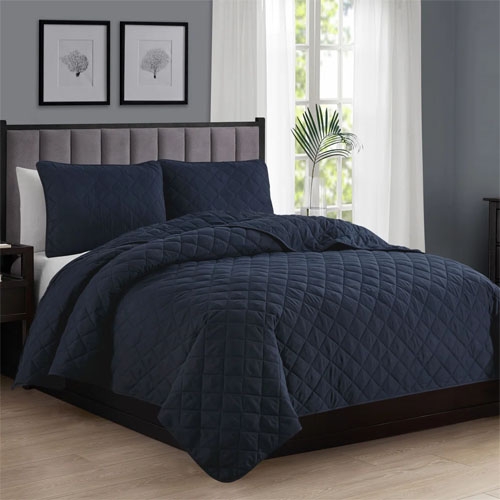 A neatly made bed with a dark blue, quilted comforter set, featuring matching pillow shams, is placed in a room with gray walls, framed art, and a window with sheer curtains.