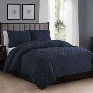 A neatly made bed with a dark blue, quilted comforter set, featuring matching pillow shams, is placed in a room with gray walls, framed art, and a window with sheer curtains.