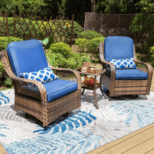 Two outdoor wicker swivel chairs with blue cushions and geometric accent pillows are arranged on a patterned outdoor rug. A small glass-top table between the chairs holds drinks and snacks, set against a green garden backdrop with decorative wooden fencing.