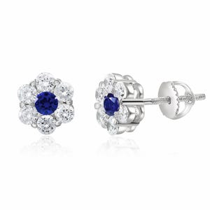Silver stud earrings with a flower design feature a blue center stone, resembling a sapphire, surrounded by smaller clear stones, resembling diamonds.