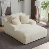 A plush, ribbed cream chaise lounge with matching pillows sits on a gray carpet, surrounded by a floor lamp, curtains, and a potted plant, creating a cozy living space.