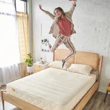 A person joyfully jumps on a minimalist wooden bed with a natural mattress. The room features a white brick wall, a patterned curtain, plants, and a small bedside table.