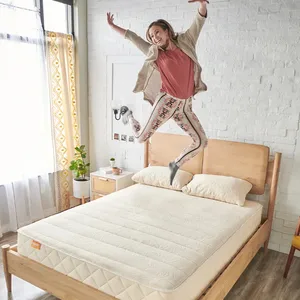 A person joyfully jumps on a minimalist wooden bed with a natural mattress. The room features a white brick wall, a patterned curtain, plants, and a small bedside table.