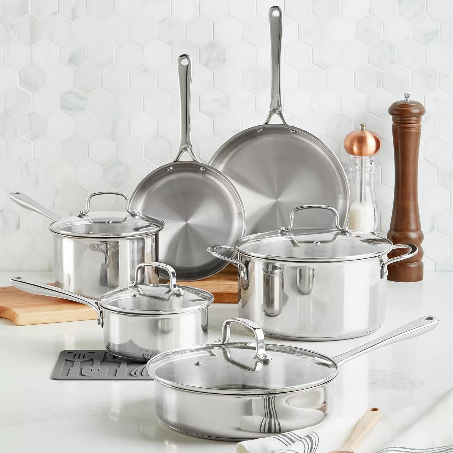 Stainless steel cookware set with two frying pans, three lidded saucepans, a salt and pepper grinder, set against a hexagonal tile backsplash.