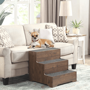 A small dog sits on a sofa next to a wooden pet staircase with three steps, featuring non-slip treads. The room includes neutral-colored decor, a lamp, plant, and patterned cushions.