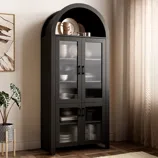 A tall, black cabinet with glass doors and a curved top section displays dinnerware, set against a neutral wall with a potted plant and curtains nearby.