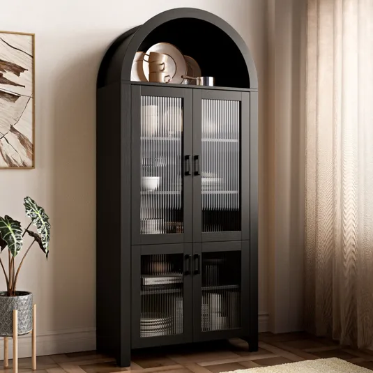 A tall, black cabinet with glass doors and a curved top section displays dinnerware, set against a neutral wall with a potted plant and curtains nearby.
