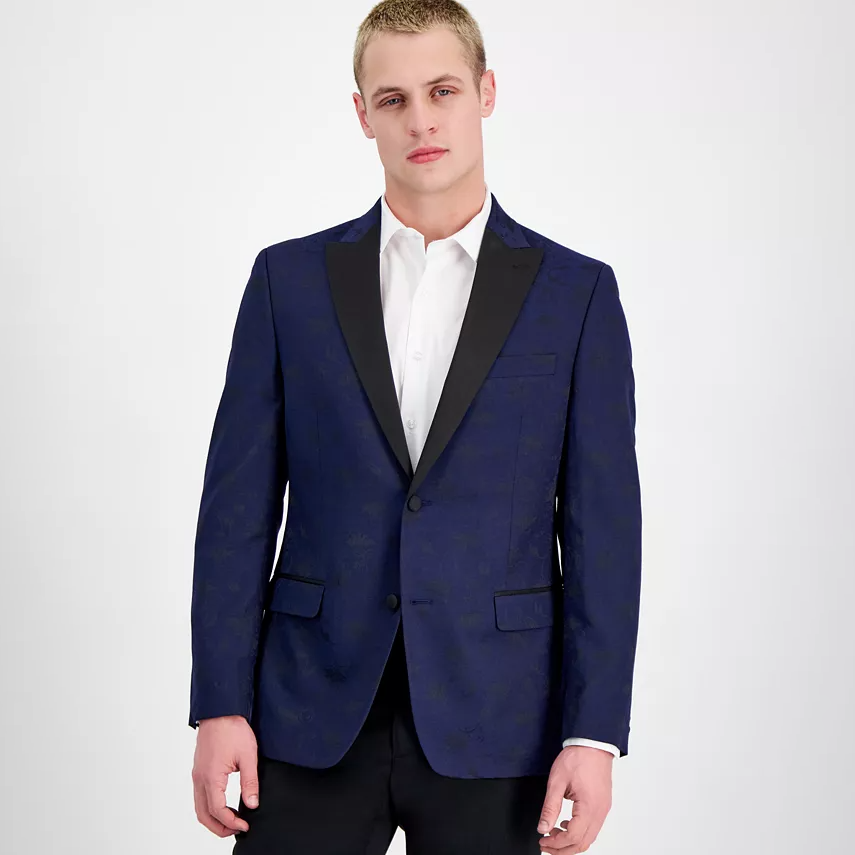 A man is wearing a navy blue, slim-fit evening jacket with a subtle floral pattern, featuring a black shawl lapel and matching pocket trims.