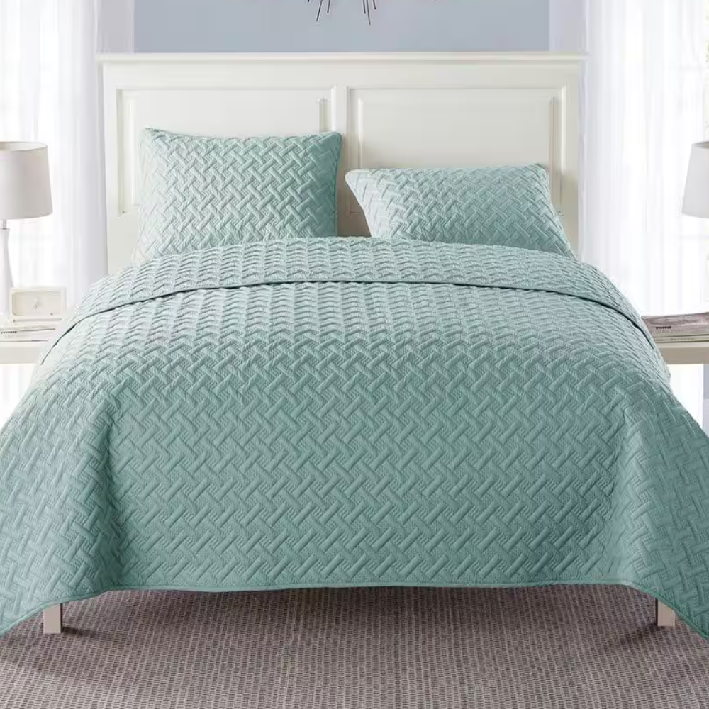 A light blue quilted bedspread with a textured pattern is shown on a bed, accompanied by two matching pillows. The bed is set against a white headboard with two bedside tables and lamps.