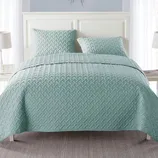 A light blue quilted bedspread with a textured pattern is shown on a bed, accompanied by two matching pillows. The bed is set against a white headboard with two bedside tables and lamps.