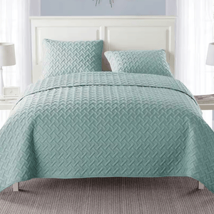 A light blue quilted bedspread with a textured pattern is shown on a bed, accompanied by two matching pillows. The bed is set against a white headboard with two bedside tables and lamps.