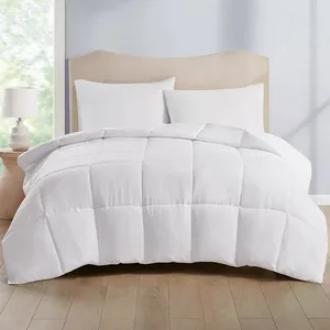 A white, quilted comforter is displayed on a bed with two matching pillows, accompanied by a light-colored headboard and a bedside table with a lamp. The setup is in a softly lit room with hardwood flooring.