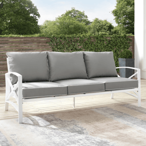 A 79'' cushioned patio sofa with a sleek white steel frame, featuring three plush gray cushions. It's set on a wooden deck with a backdrop of greenery, creating an elegant outdoor seating option.