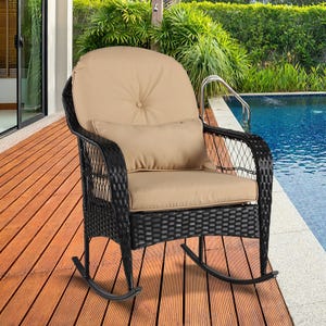 A black wicker rocking chair with beige cushions is set on a wooden deck beside a pool, with lush greenery in the background.