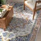 A decorative rug with intricate geometric and floral patterns in blue, brown, and green tones is placed under a wooden coffee table and next to a cushioned chair with a book on the seat. A cup of coffee and a plant adorn the table.