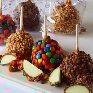 A variety of Mrs. Prindables candy apples, including ones coated in colorful candy, nuts, and chocolate chips. Cut slices of green apple surround the whole apples, which are individually wrapped in clear packaging.