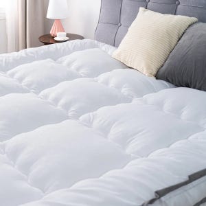 4\“ Queen Down Alternative Mattress Topper features a soft, quilted surface for added comfort. It is shown on a bed with a tufted headboard, accompanied by a white lamp on a nightstand and pillows in cream and gray tones.