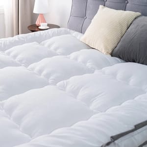 4\“ Queen Down Alternative Mattress Topper features a soft, quilted surface for added comfort. It is shown on a bed with a tufted headboard, accompanied by a white lamp on a nightstand and pillows in cream and gray tones.