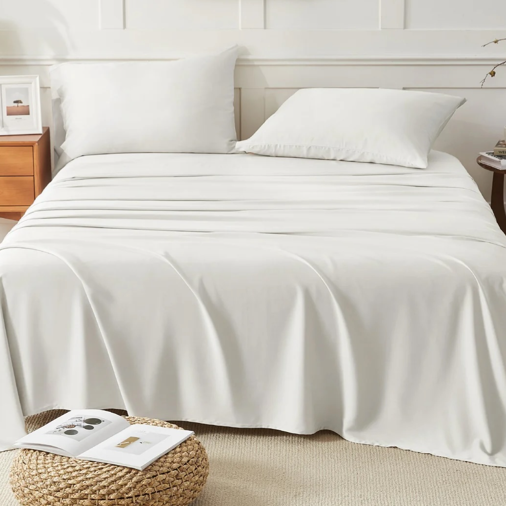 A neatly made bed with white sheets and pillows is flanked by a wooden nightstand with a lamp and photo frame on the left and a small round table with books and a plant on the right. A woven stool with an open book is in front.