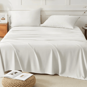 A neatly made bed with white sheets and pillows is flanked by a wooden nightstand with a lamp and photo frame on the left and a small round table with books and a plant on the right. A woven stool with an open book is in front.