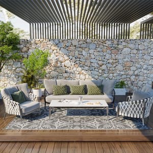 Patio set featuring a cushioned sofa, two matching chairs, a rectangular coffee table, and decor pillows on a patterned outdoor rug, set against a stone wall background with greenery.