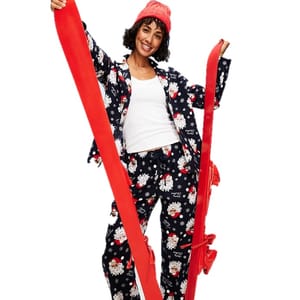 A person wearing a matching pajama set with a Santa theme, a white tank top, and a red knit hat is holding two large red skis.