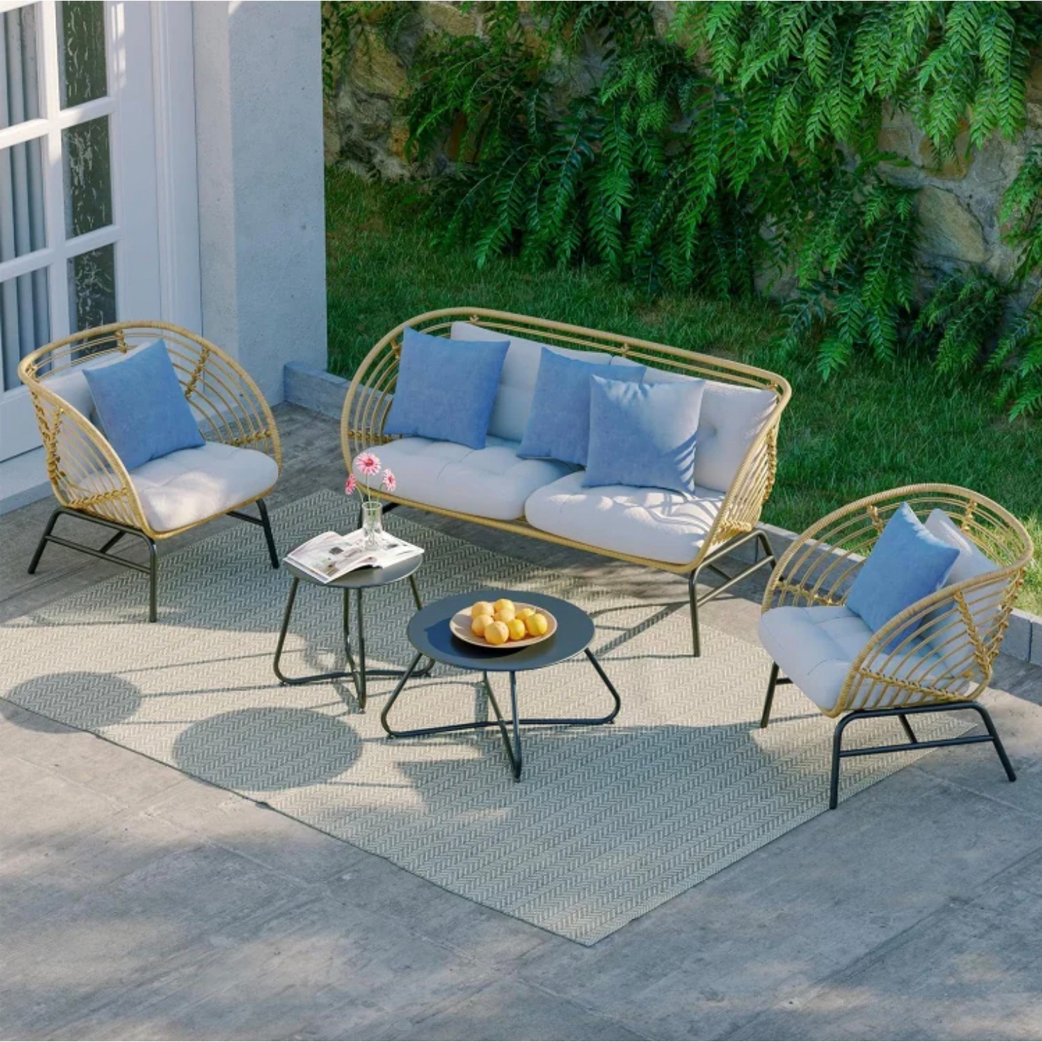 A five-piece patio set includes a wicker-style loveseat and two chairs with cream cushions and blue pillows, accompanied by two round black side tables placed on a light-patterned outdoor rug. The setting is adjacent to a garden with greenery.