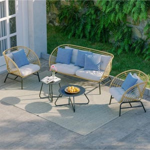A five-piece patio set includes a wicker-style loveseat and two chairs with cream cushions and blue pillows, accompanied by two round black side tables placed on a light-patterned outdoor rug. The setting is adjacent to a garden with greenery.