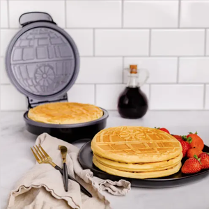 A waffle maker designed to create waffles with a Death Star pattern, accompanied by a plate of waffles and strawberries, and a syrup bottle in the background.