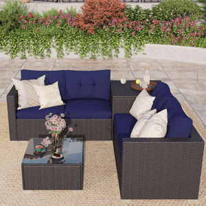 Outdoor patio set with navy cushions includes an L-shaped sectional sofa and a glass-top coffee table, placed on a woven rug, with decorative pillows and flowers, set against a backdrop of lush greenery.
