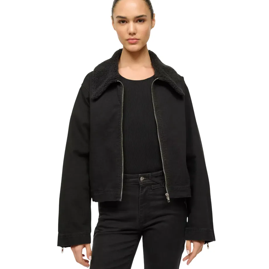 A person is wearing a black jacket with a sherpa collar over a black ribbed shirt and matching black pants.