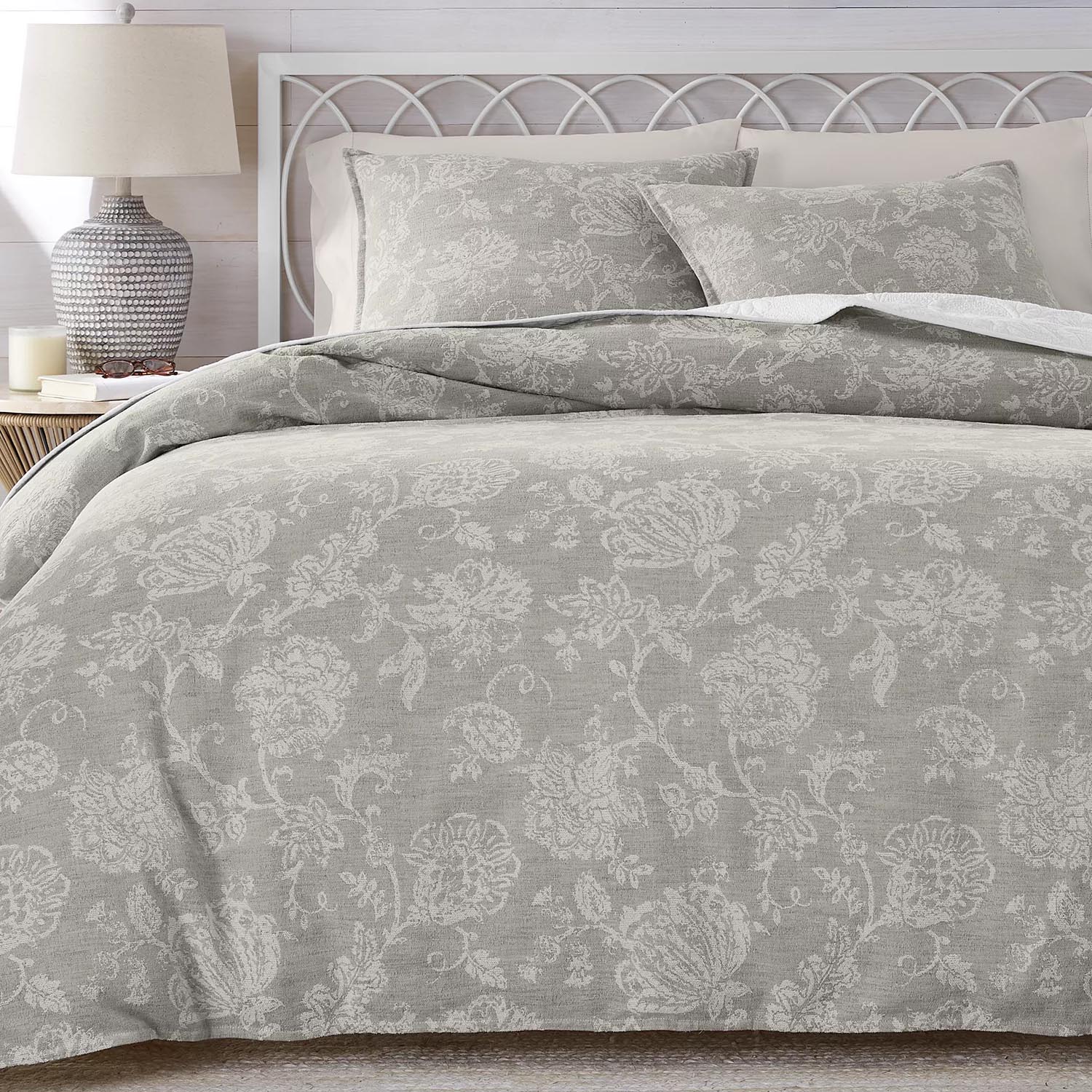 A bed features a grey floral-patterned comforter set with matching pillow shams. A bedside table holds a textured lamp, books, and a candle.