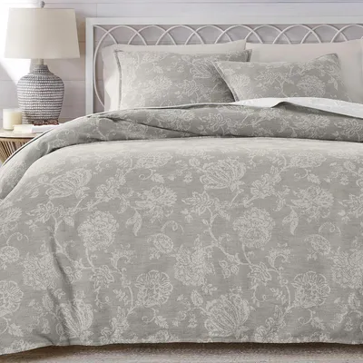 A bed features a grey floral-patterned comforter set with matching pillow shams. A bedside table holds a textured lamp, books, and a candle.