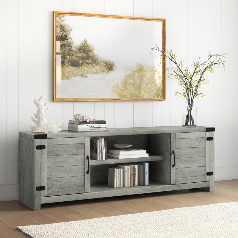 A 70\“ TV stand with a rustic gray finish, featuring two cabinet doors with black metal handles, open shelving for storage, and black metal corner accents. It's decorated with books, a vase with branches, and coral, set against a paneled wall and framed artwork.