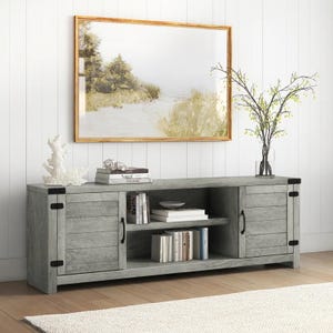 A 70\“ TV stand with a rustic gray finish, featuring two cabinet doors with black metal handles, open shelving for storage, and black metal corner accents. It's decorated with books, a vase with branches, and coral, set against a paneled wall and framed artwork.
