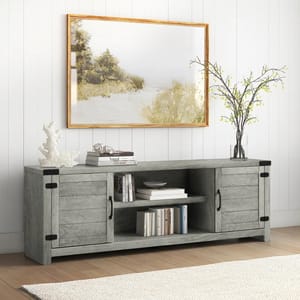 A 70\“ TV stand with a rustic gray finish, featuring two cabinet doors with black metal handles, open shelving for storage, and black metal corner accents. It's decorated with books, a vase with branches, and coral, set against a paneled wall and framed artwork.