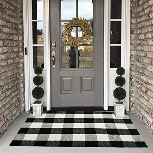 A black and white buffalo plaid rug is laid out on a porch, flanked by topiary trees in planters and positioned in front of a door with a wreath.