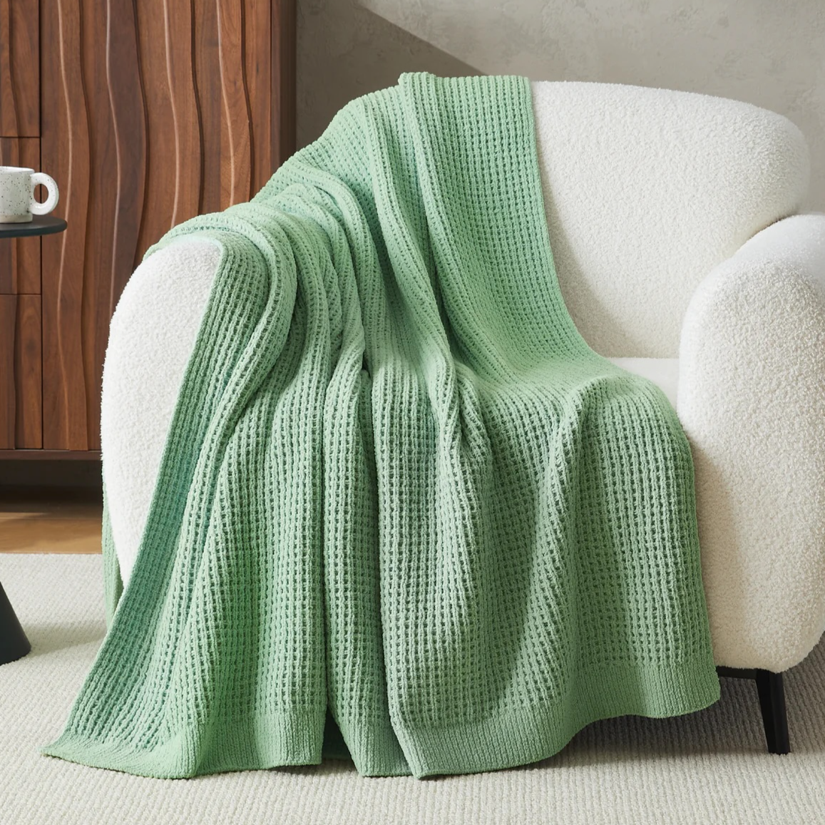 A soft, white boucle armchair draped with a textured green knit throw blanket, positioned in a cozy room with a wooden cabinet and round black side table, alongside a coffee cup.