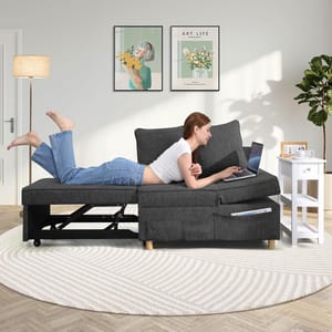 A person is lying on a dark gray futon with wooden legs, using a laptop; a white side table and a floor lamp are also visible.